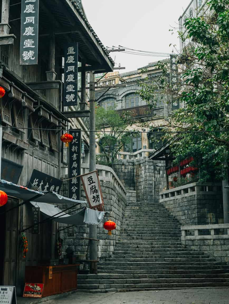 Stone steps leading up between traditional buildings with hanging red lanterns and signs, surrounded by greenery under an overcast sky.