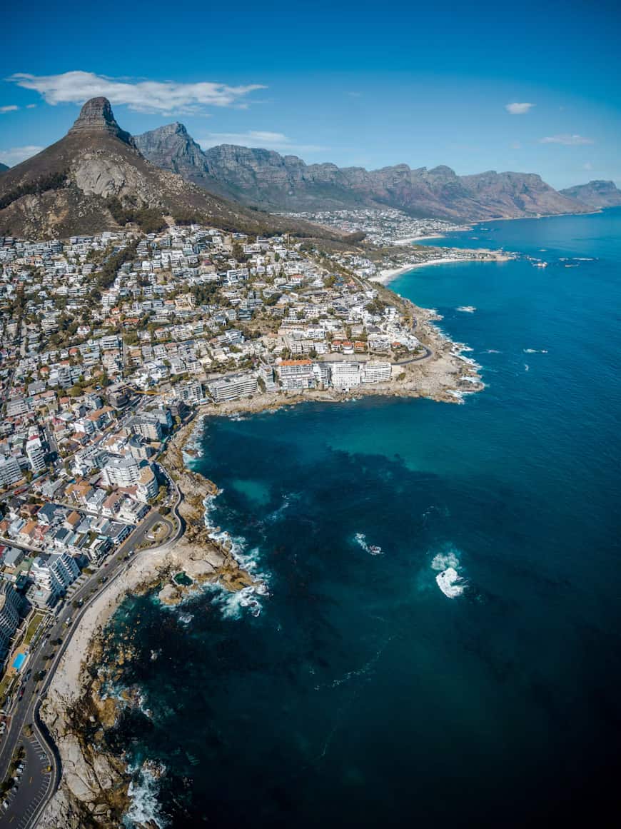 Aerial view of a coastal city with rugged mountains, vibrant blue ocean, white buildings on the shoreline, and a winding coastal road.
