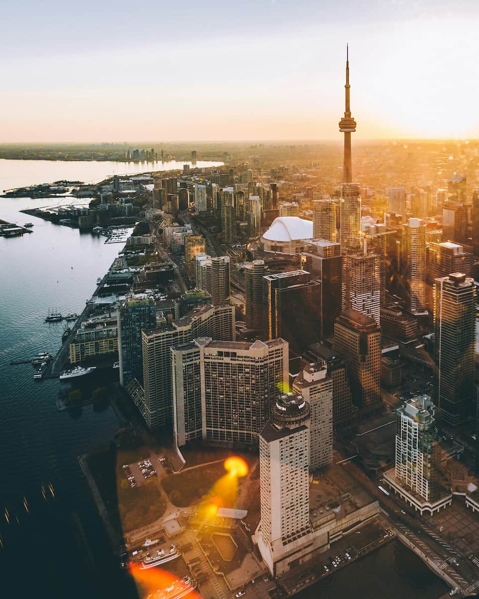 Aerial view of a city skyline with a prominent tower at sunset, surrounded by tall buildings and water, with warm sunlight illuminating the scene.