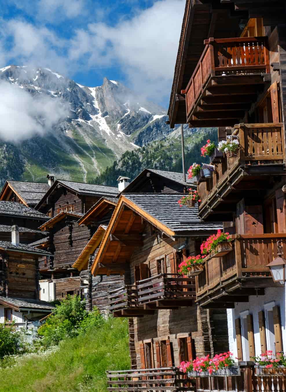 Wooden alpine houses with flowered balconies, set on a grassy slope with snow-capped mountains and a partly cloudy blue sky in the background.