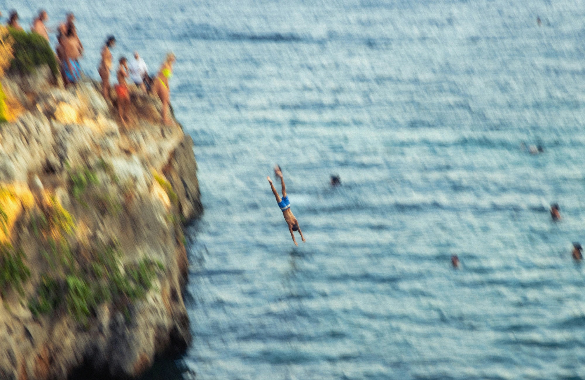 A cliff diver mid-dive towards the sea while friends watch from the rocks
