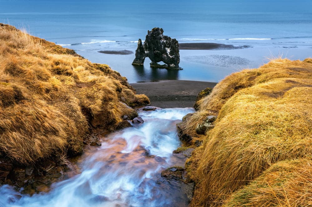 Scenic landscape featuring a flowing stream between grassy hills leading to a dramatic rock formation in the ocean, under a cloudy sky