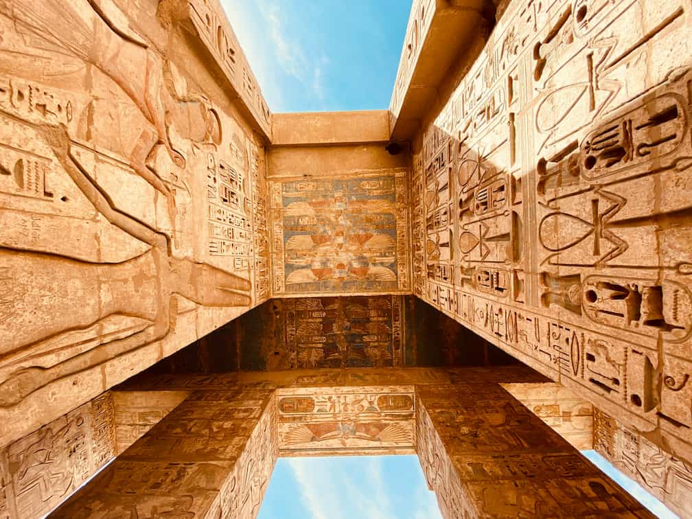 View looking up at ancient Egyptian temple walls with detailed hieroglyphics and carvings, set against a clear blue sky.