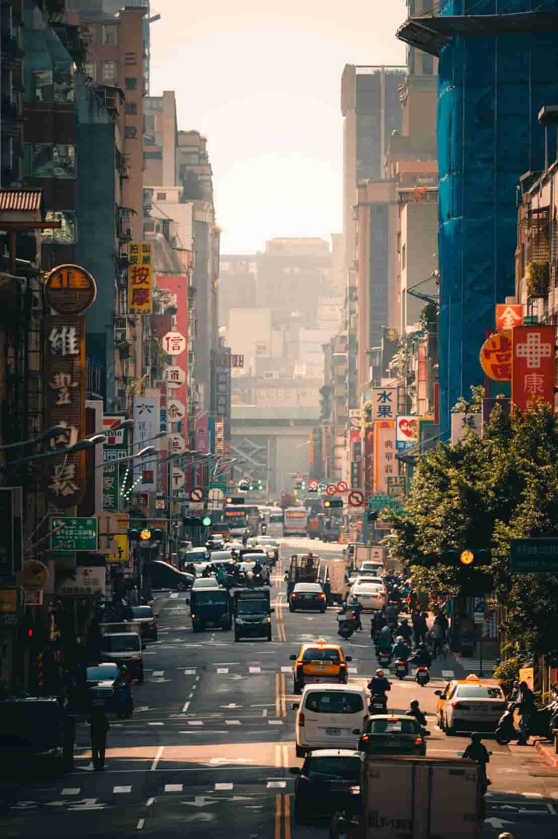 Busy urban street with heavy traffic, colorful signage in Chinese, and tall buildings on both sides under a hazy sky.