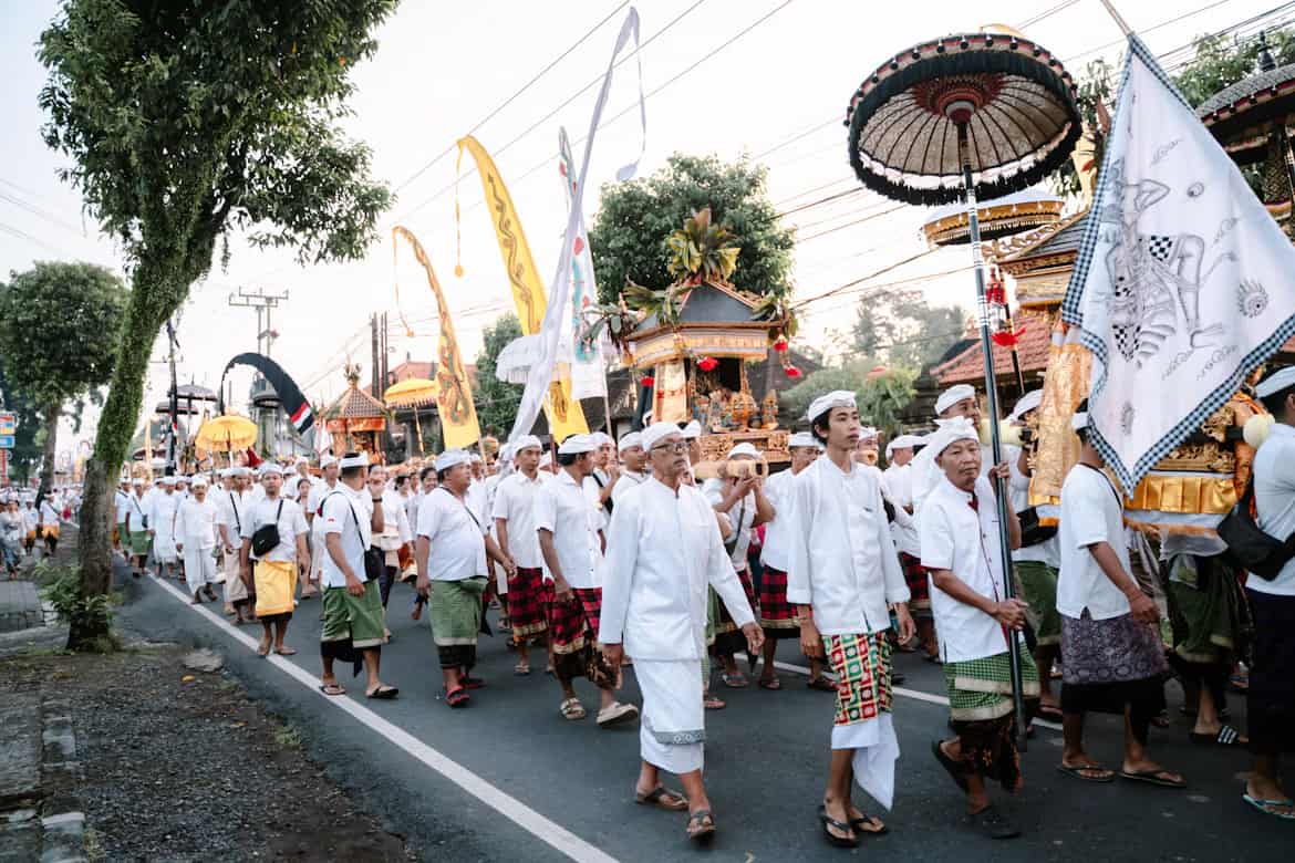 A vibrant procession in traditional attire walks along a street with colorful banners and ceremonial decorations.