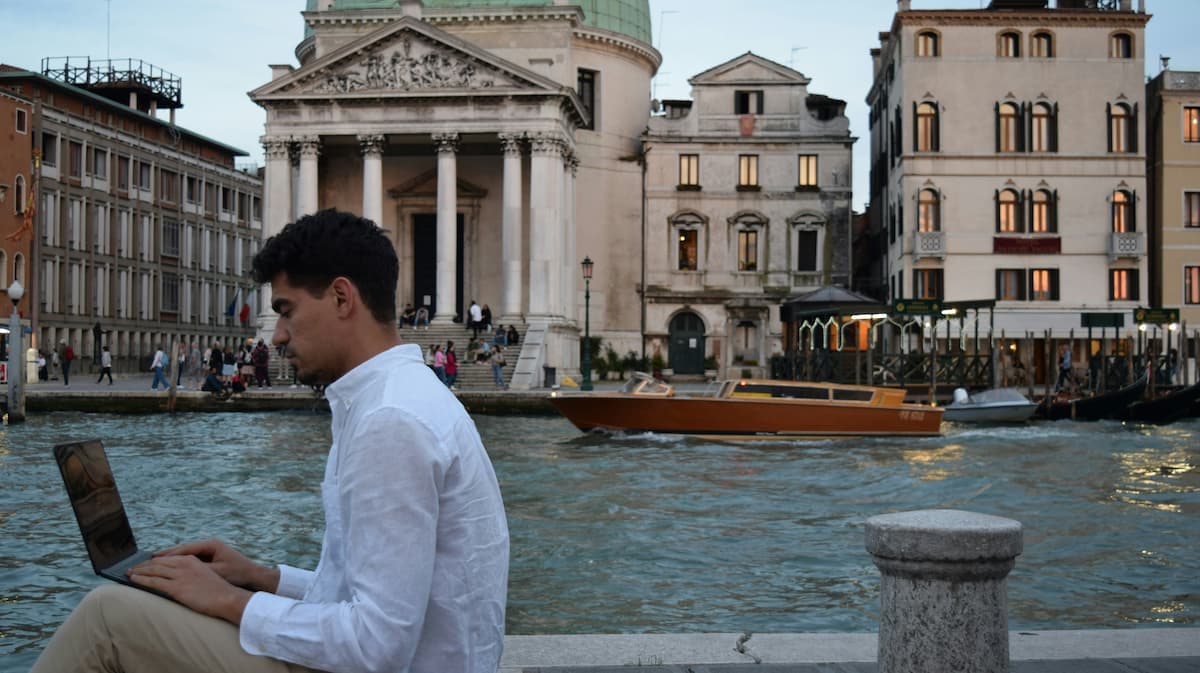 A man in a white shirt sits by a canal with a laptop, with elegant buildings and a boat on the water in the background.