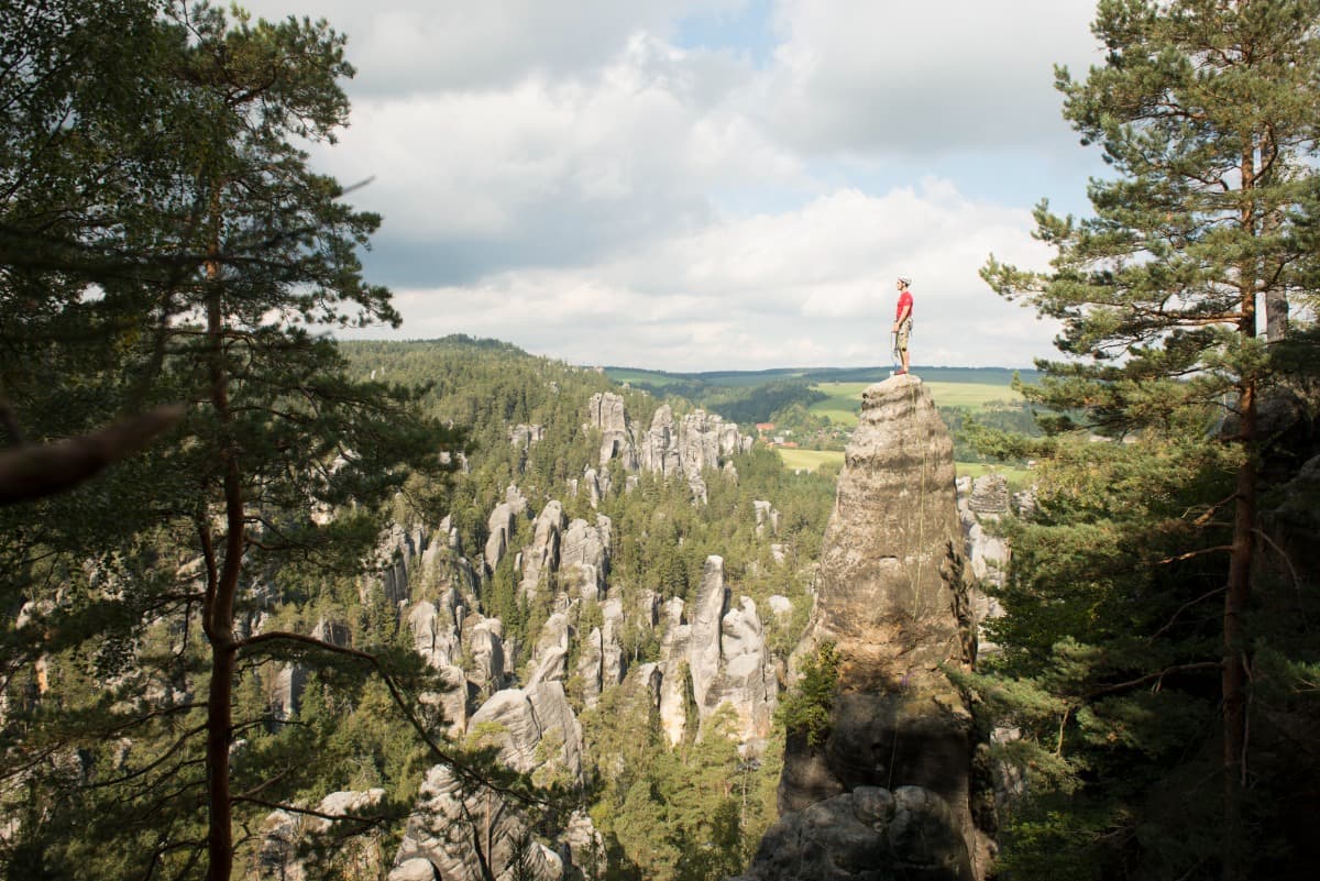 Person climbing a tall rock formation, surrounded by a forested landscape with scattered rocky spires under a partly cloudy sky.