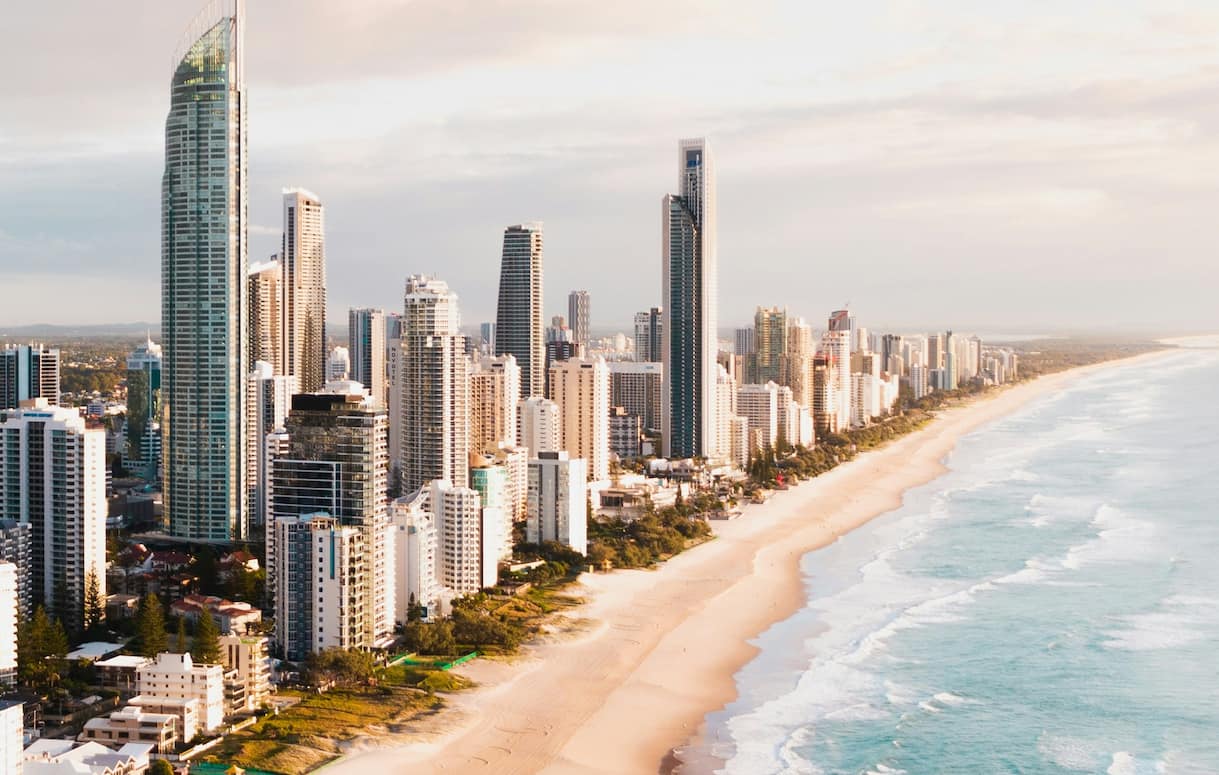 Aerial view of a city skyline with tall buildings along a sandy beach and ocean waves under a cloudy sky.