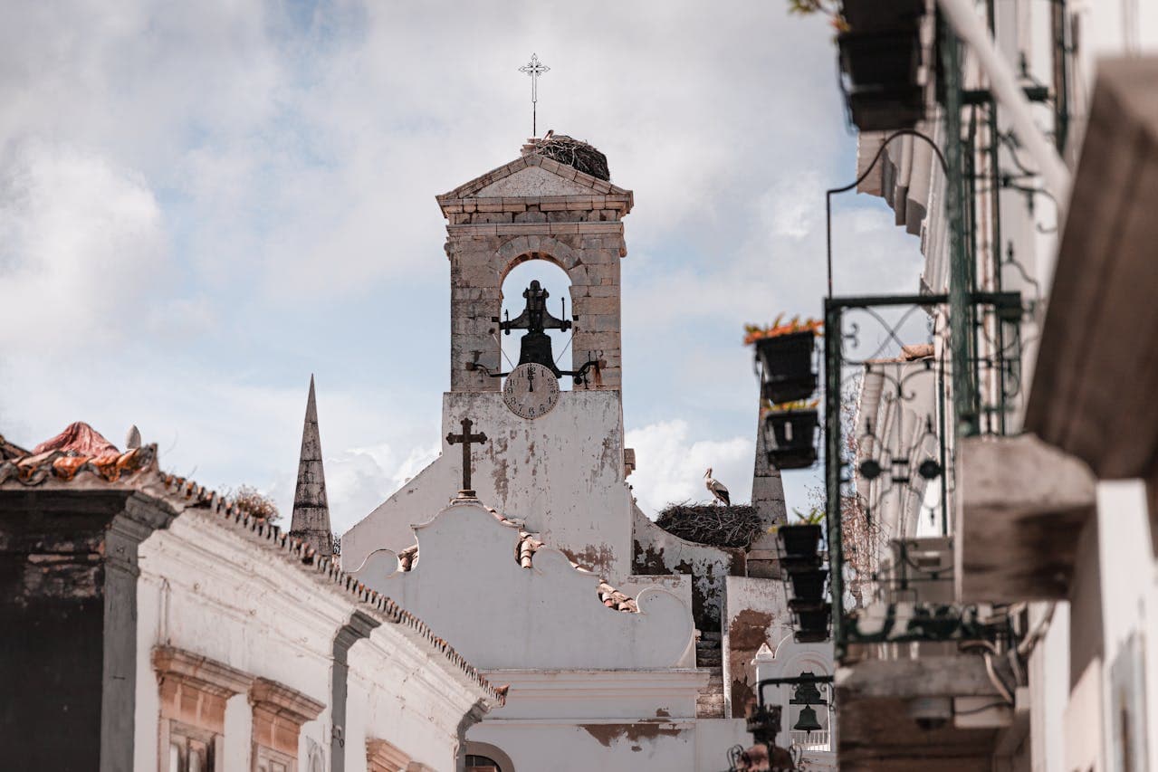 Sé Cathedral, Faro. Old whitewashed church with a bell tower and clock, topped by a cross and a stork’s nest, seen from a narrow street with hanging flower baskets.