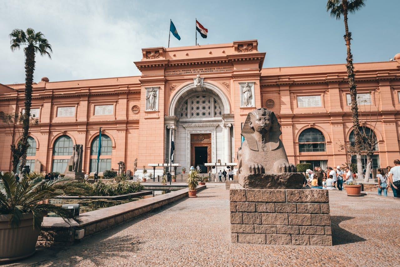 Granite sphinx statue in front of the Egyptian Museum in Cairo, surrounded by visitors and palm trees.
