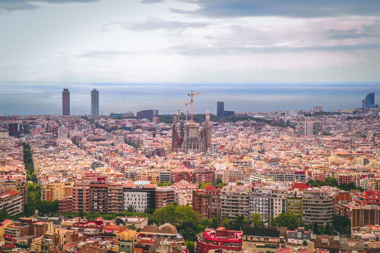 A panoramic view of Barcelona, Spain, featuring the iconic Sagrada Família basilica under construction in the center, with the Mediterranean Sea and city skyline in the background.