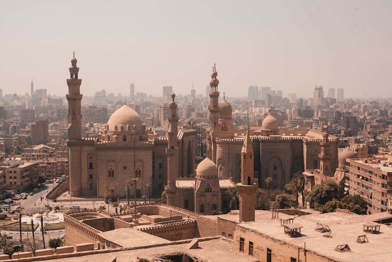 Panoramic view of the Cairo skyline showcasing the historic Sultan Hassan Mosque and Al-Rifa'i Mosque with minarets and domes, framed by modern buildings and a hazy cityscape.
