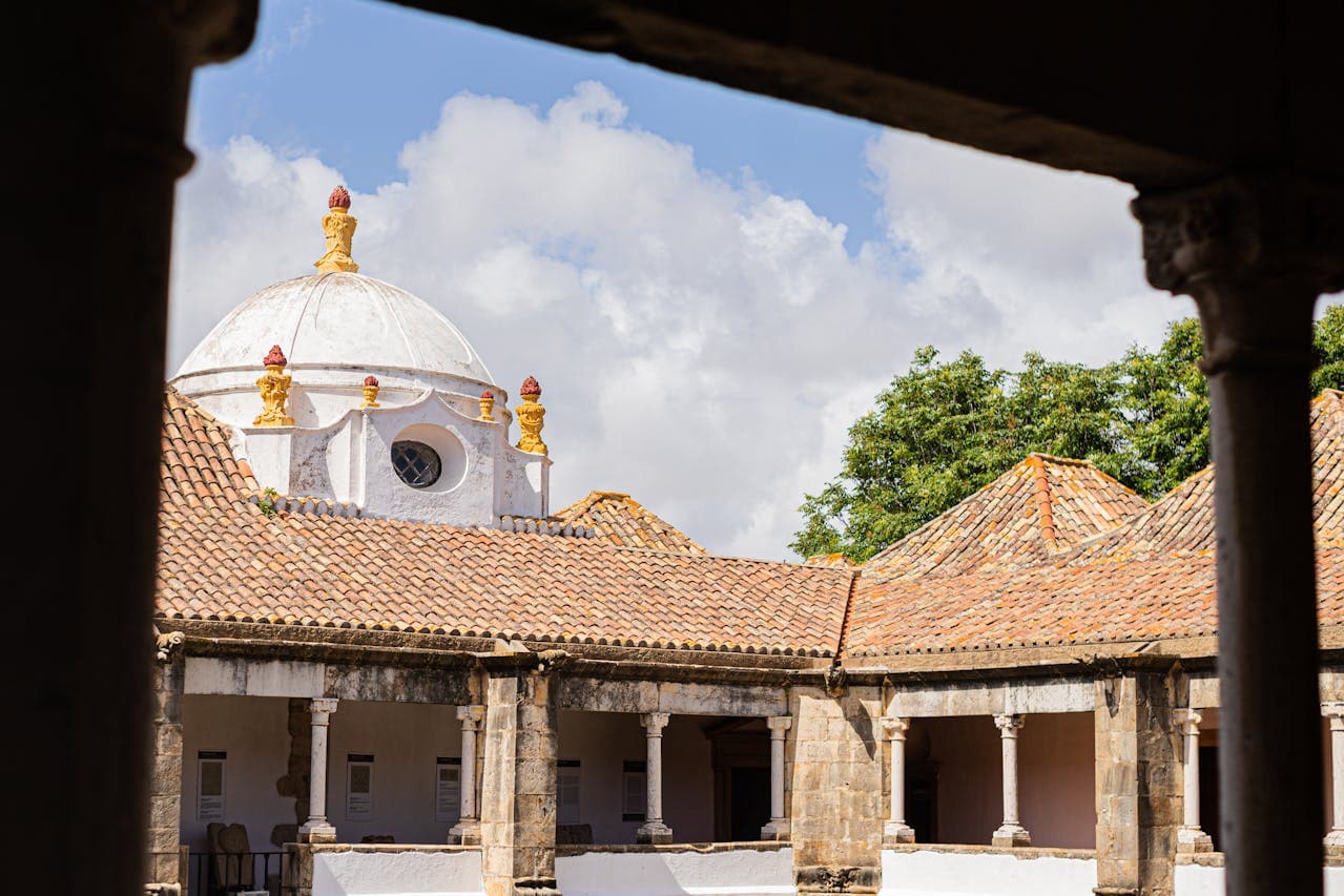 The view from a shaded cloister looking out onto a historic building with a white domed roof, ornate yellow and red decorations, and terracotta tiles, surrounded by stone columns and lush green trees.