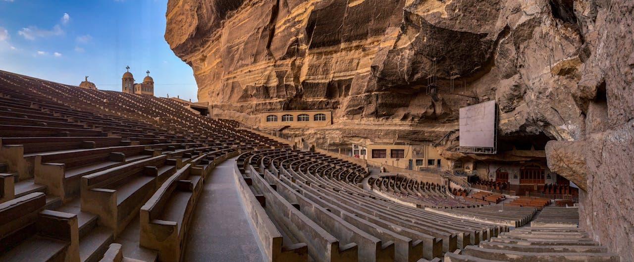 Wide-angle view of a large amphitheater carved into a rocky cliff, with rows of stone seating curving around a central stage area. The structure is partially sheltered by the overhanging rock, and church domes with crosses are visible in the background against a blue sky
