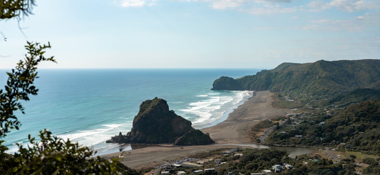 Scenic aerial view of Piha Beach on New Zealand’s west coast, highlighting Lion Rock, black sand shoreline, blue ocean waves, and lush green hills in the background.