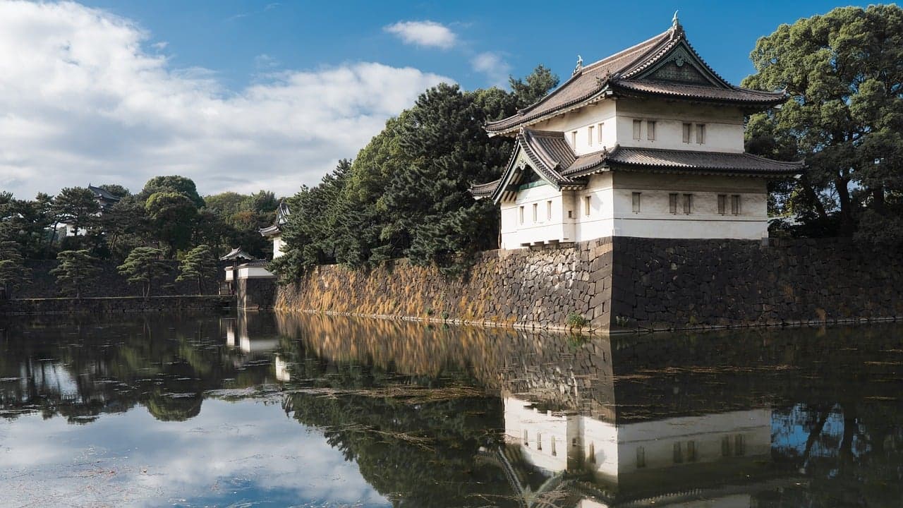 White traditional Japanese castle turret on a stone wall beside a calm moat reflecting the building, trees and blue sky.