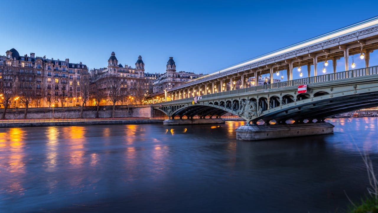 A green iron bridge spanning a river in a European city, with historic buildings and dramatic clouds in the background.