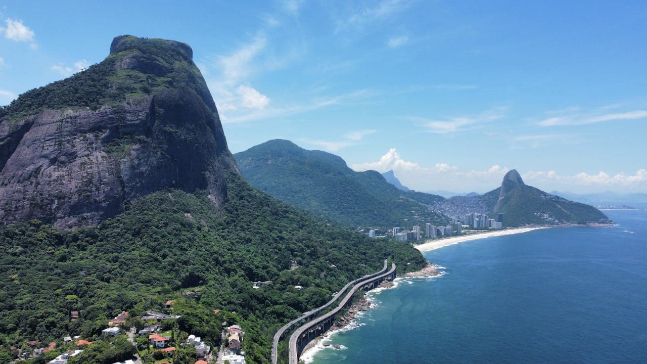 Aerial view of Rio de Janeiro’s coastline with lush green mountains, a winding coastal road, and the iconic Pedra da Gávea rock formation under a clear blue sky.