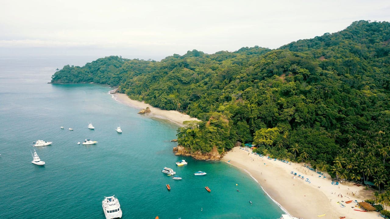 Tropical beach with turquoise waters, anchored boats, and a forested coastline, showcasing a popular tourist destination in Costa Rica for boating and relaxation.