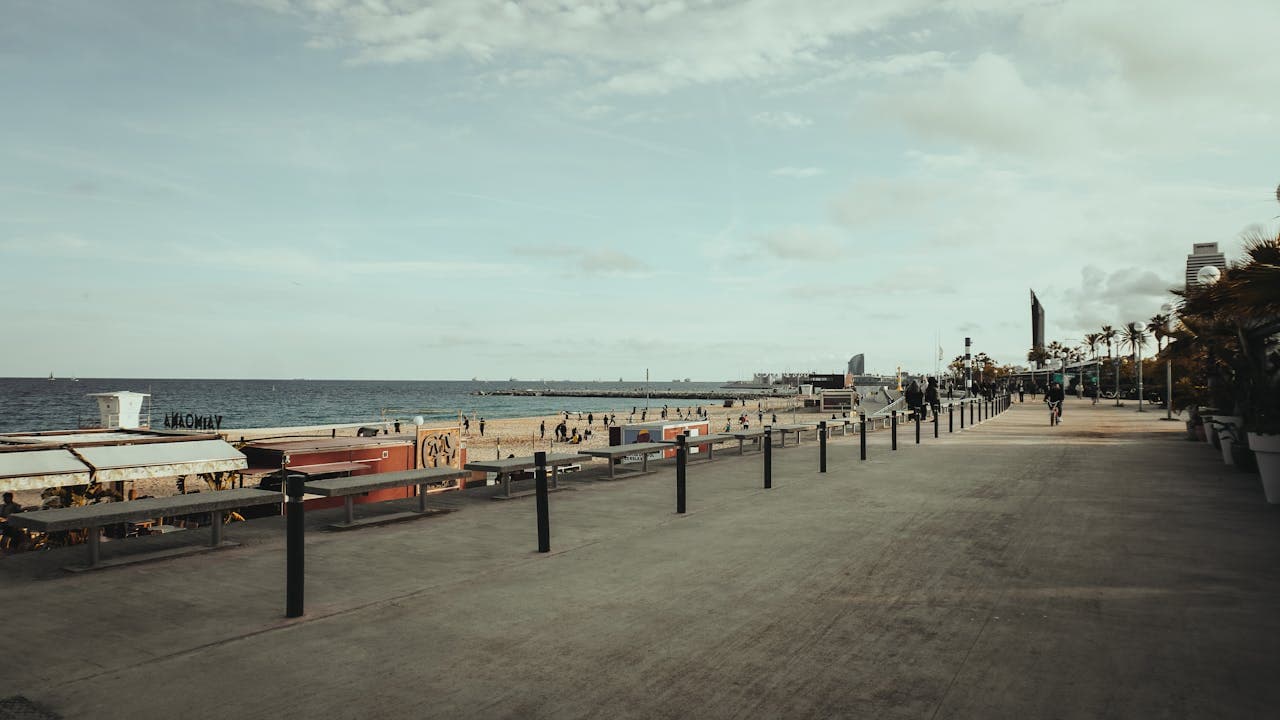 Wide promenade and sandy beach with people walking and playing near the sea in Barcelona, Spain, with palm trees and cityscape in the background.