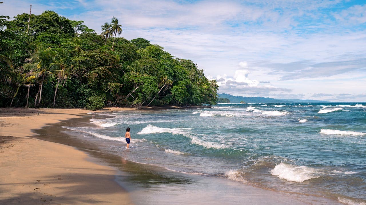 Woman standing on a secluded tropical beach with lush green jungle, blue ocean waves, and clear sky.