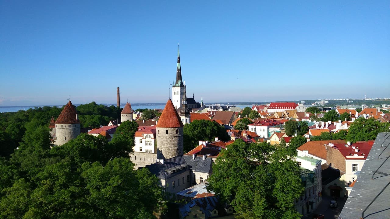 Aerial view of a picturesque town with red-roofed buildings, a tall spire, and lush green trees under a clear blue sky.