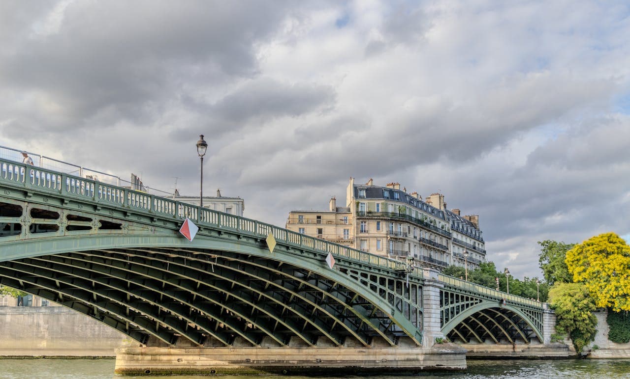 Ornate green iron bridge spanning the Seine River in Paris with classic Haussmann‑style apartment buildings under a dramatic cloudy sky