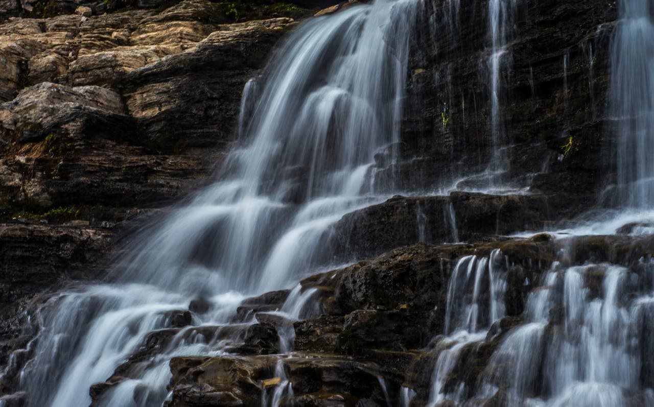A beautiful cascading waterfall flowing over dark, layered rocks in a natural setting, with the water appearing soft and misty due to long exposure photography.