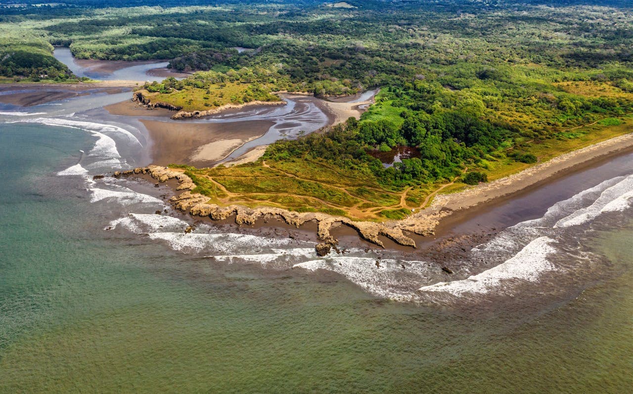 Aerial view of a scenic coastal landscape with rocky shoreline, green forest, and ocean waves in a tropical environment.