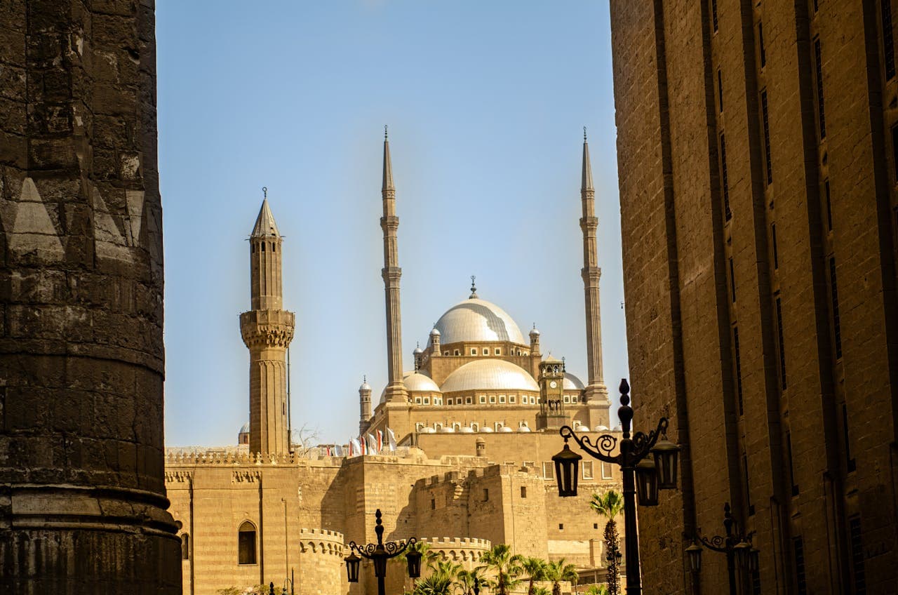 A close view of the Mosque of Muhammad Ali in Cairo, Egypt, framed between two large stone walls. The mosque's large domes and tall minarets stand prominently under a bright blue sky.