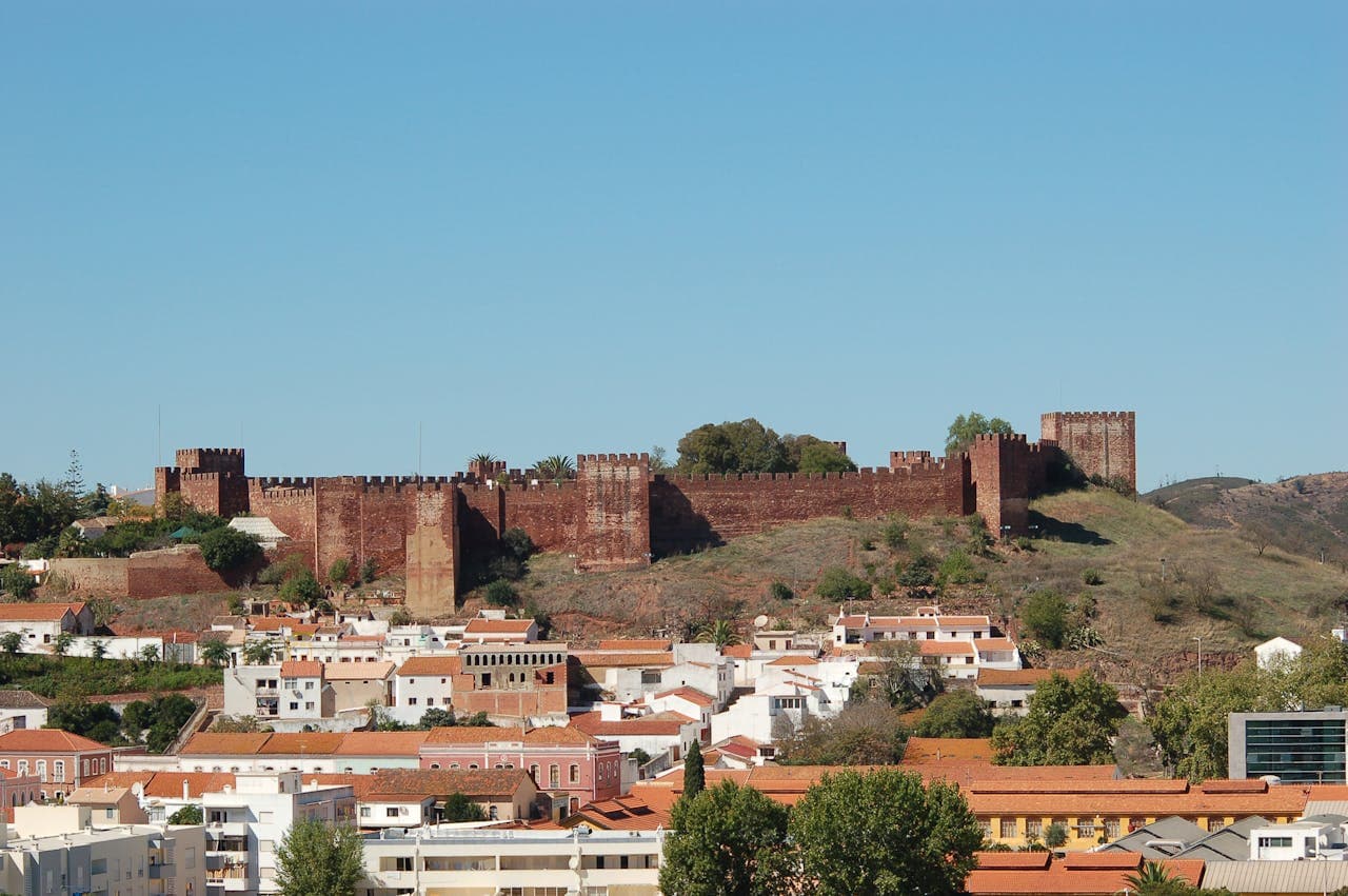 Large medieval red-brick castle with crenellated walls and towers, perched on a hill overlooking a town with white and terracotta-roofed buildings.