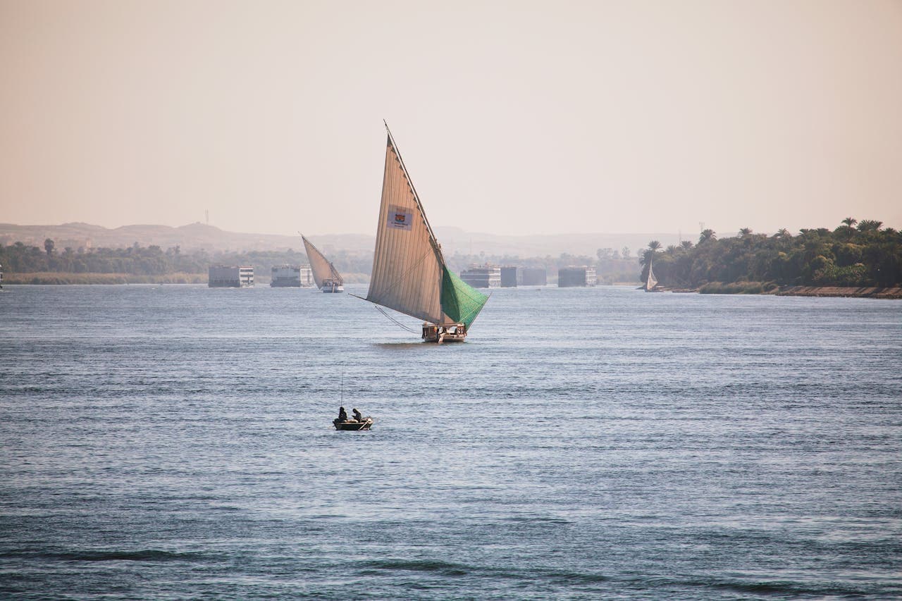 Traditional sailboats and a small fishing boat on the Nile River with green riverbanks and a hazy sky.