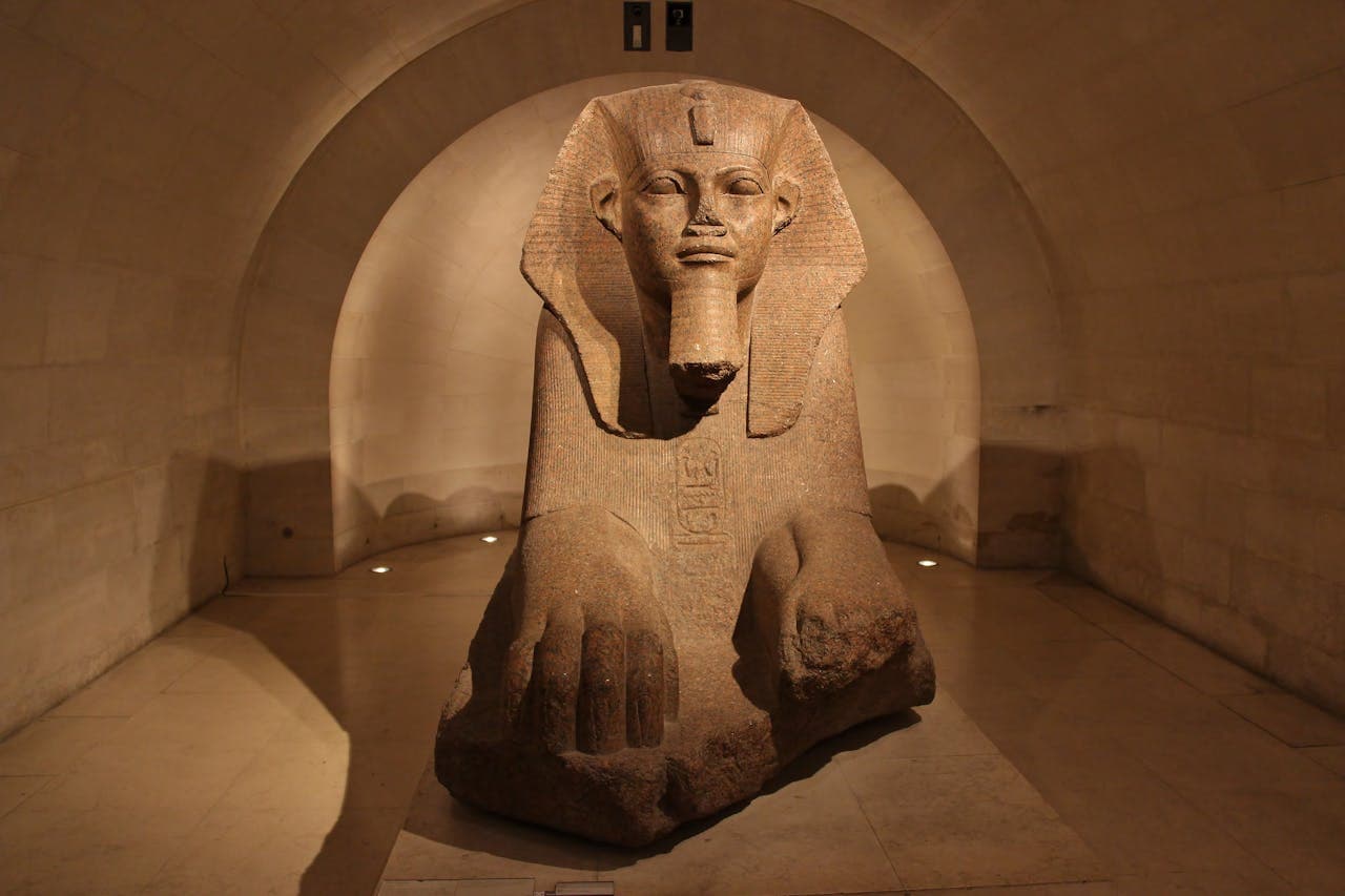 Ancient Egyptian sphinx statue displayed in a museum with arched stone walls and dramatic lighting.