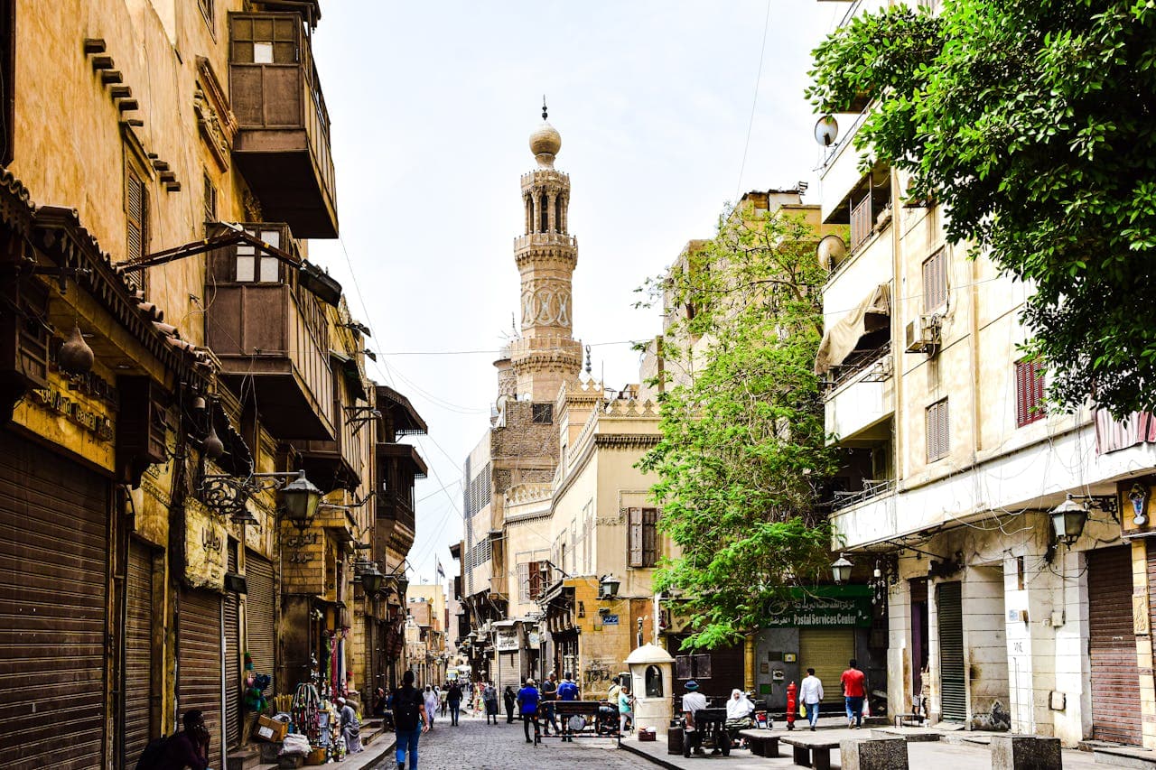 Vibrant street scene in Islamic Cairo lined with traditional wooden balconies, market stalls, and the minaret of an old mosque in the background, with people walking on cobblestone streets.