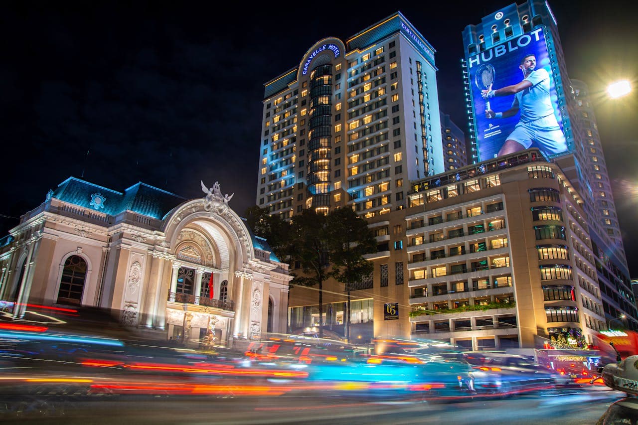 Nighttime scene of the Saigon Opera House and Caravelle Hotel in Ho Chi Minh City, Vietnam, with light trails from city traffic and illuminated buildings.