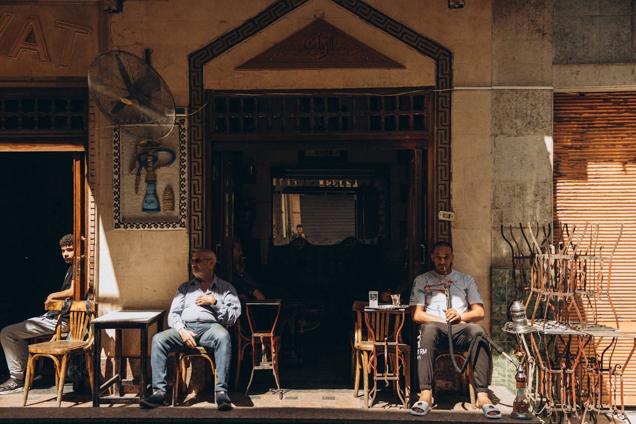 An outdoor café scene in a Middle Eastern or North African city, with two men sitting on wooden chairs outside the entrance. The café has traditional architectural details, such as decorative tiles and carved woodwork, and there are small tables with drinks and a hookah on display.