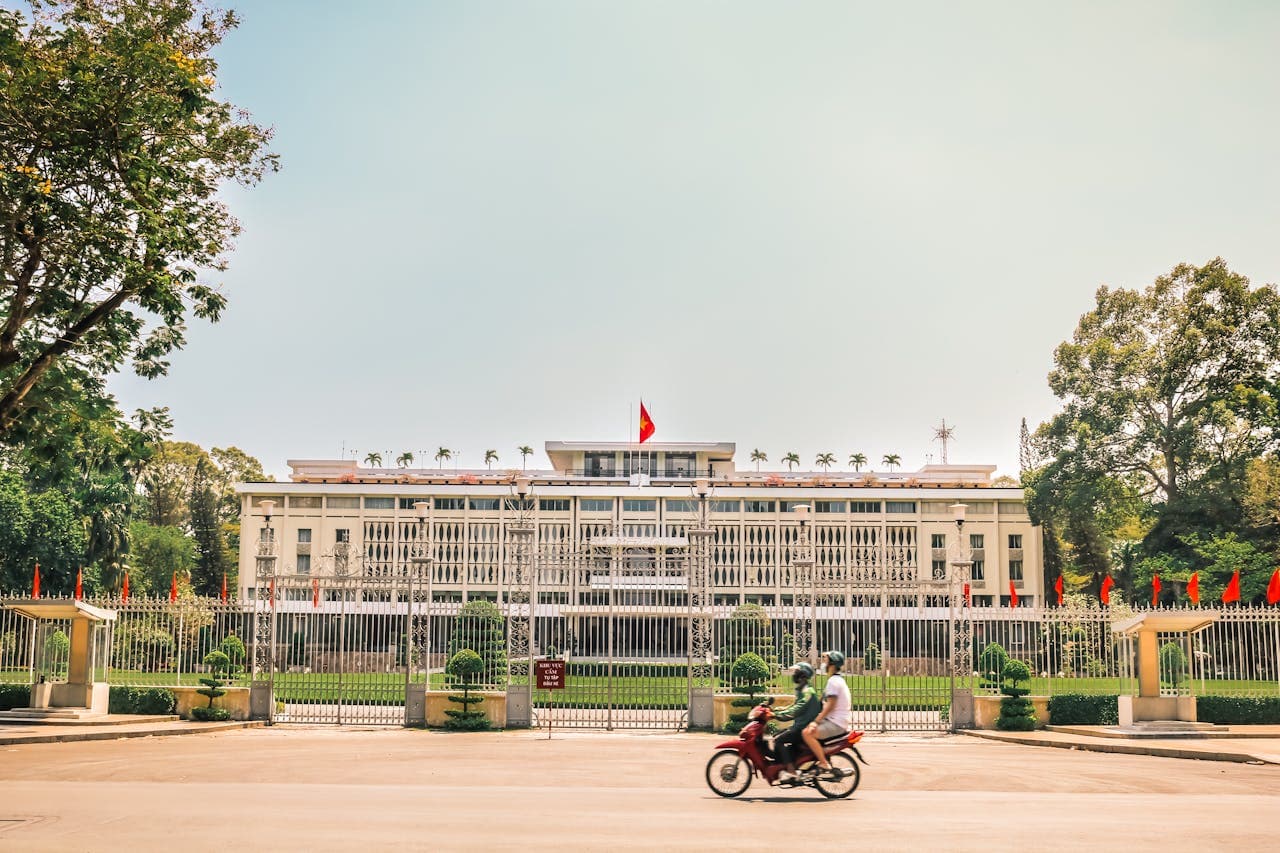 Front view of the Independence Palace (Reunification Palace) in Ho Chi Minh City, Vietnam, with the Vietnamese flag flying above and motorbikes passing by.