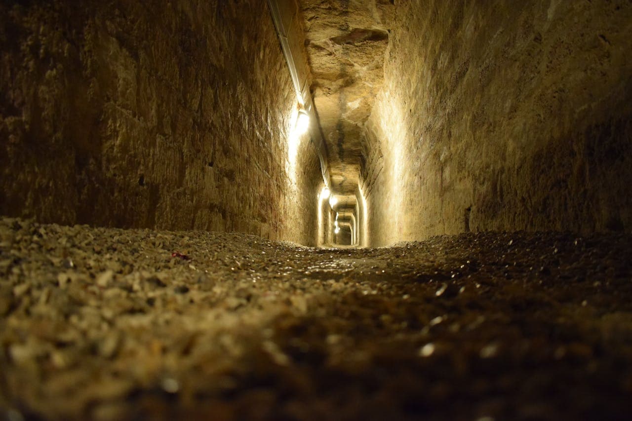 A dimly lit underground stone tunnel with a gravel floor and lights lining one wall.