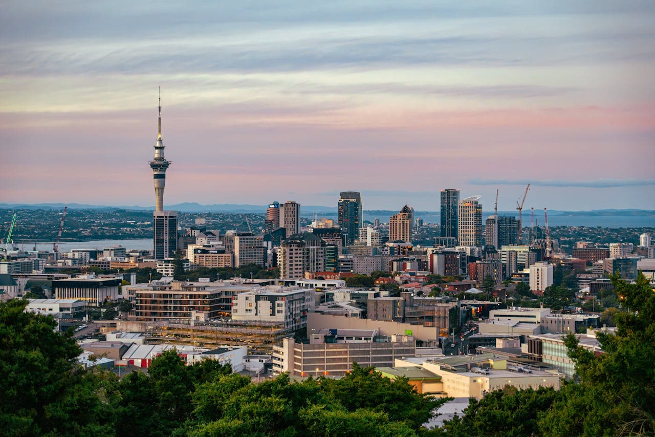 Auckland skyline at sunset with the iconic Sky Tower rising above modern high‑rise buildings and the harbour, under a pastel pink and blue sky.