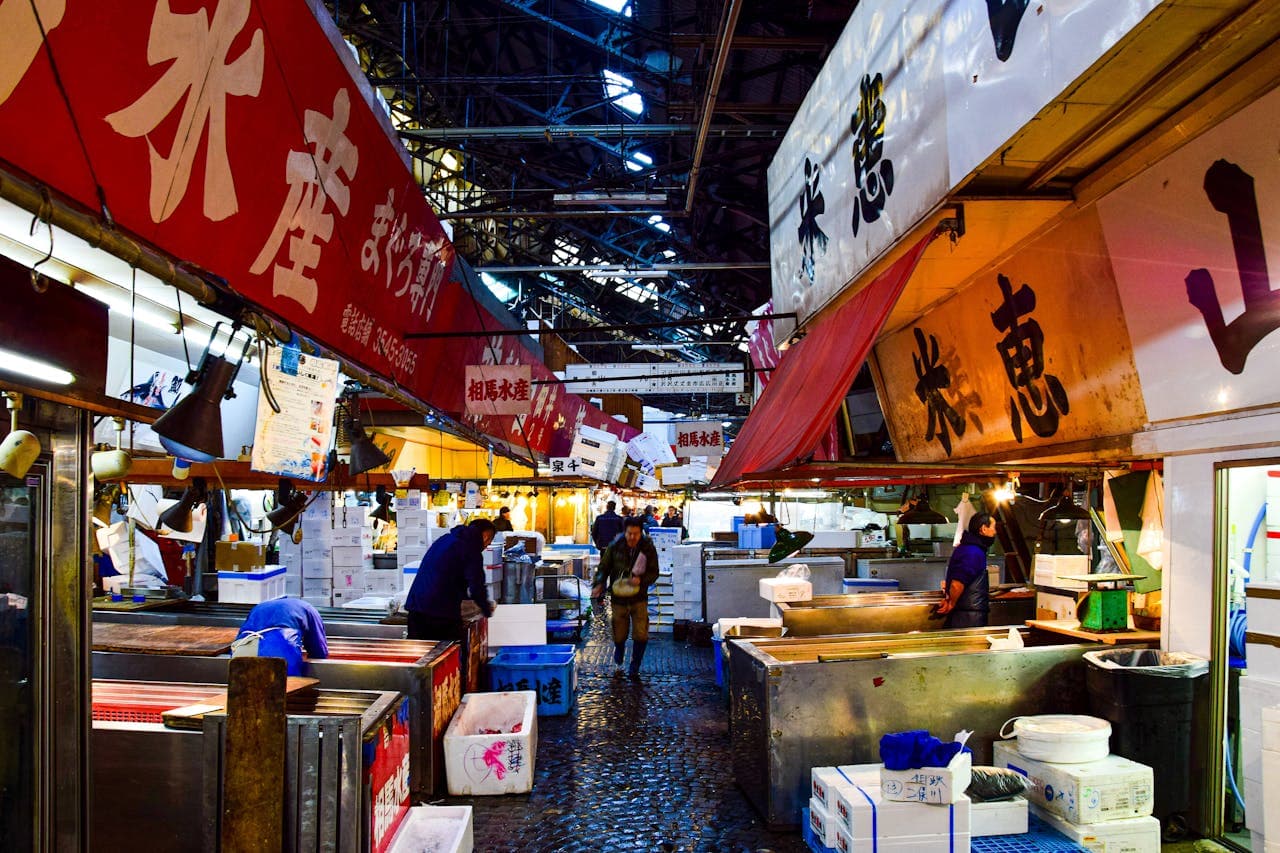 Busy indoor Japanese fish market aisle with red awnings, vendors and workers, stacked white boxes and ice bins on a wet cobblestone floor.