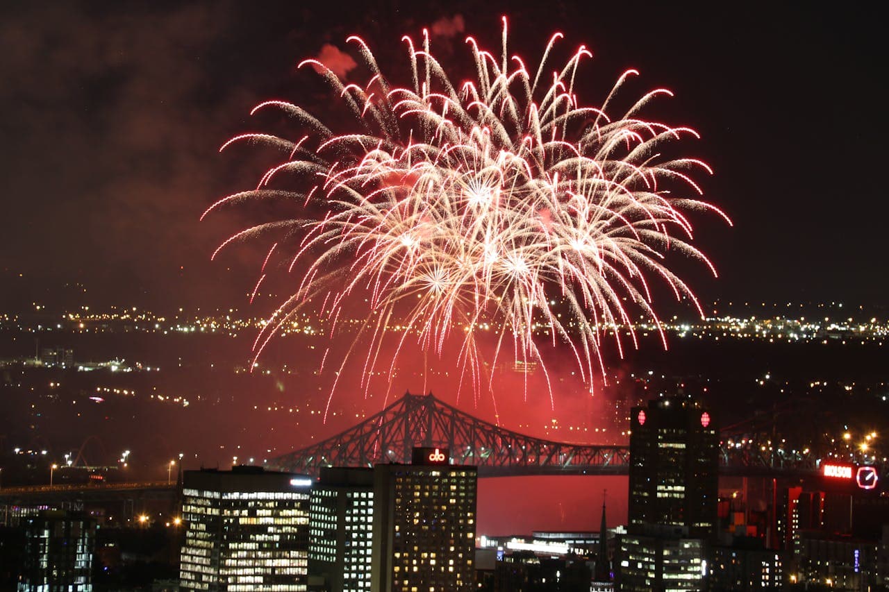 Red fireworks explode over a cityscape at night, with a bridge and illuminated buildings in the background.