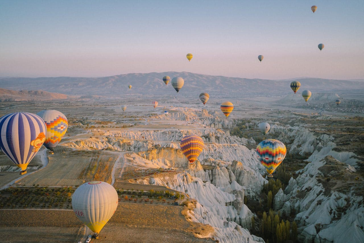Hot air balloons float over a scenic landscape with canyons and distant mountains under a clear sky at sunrise.
