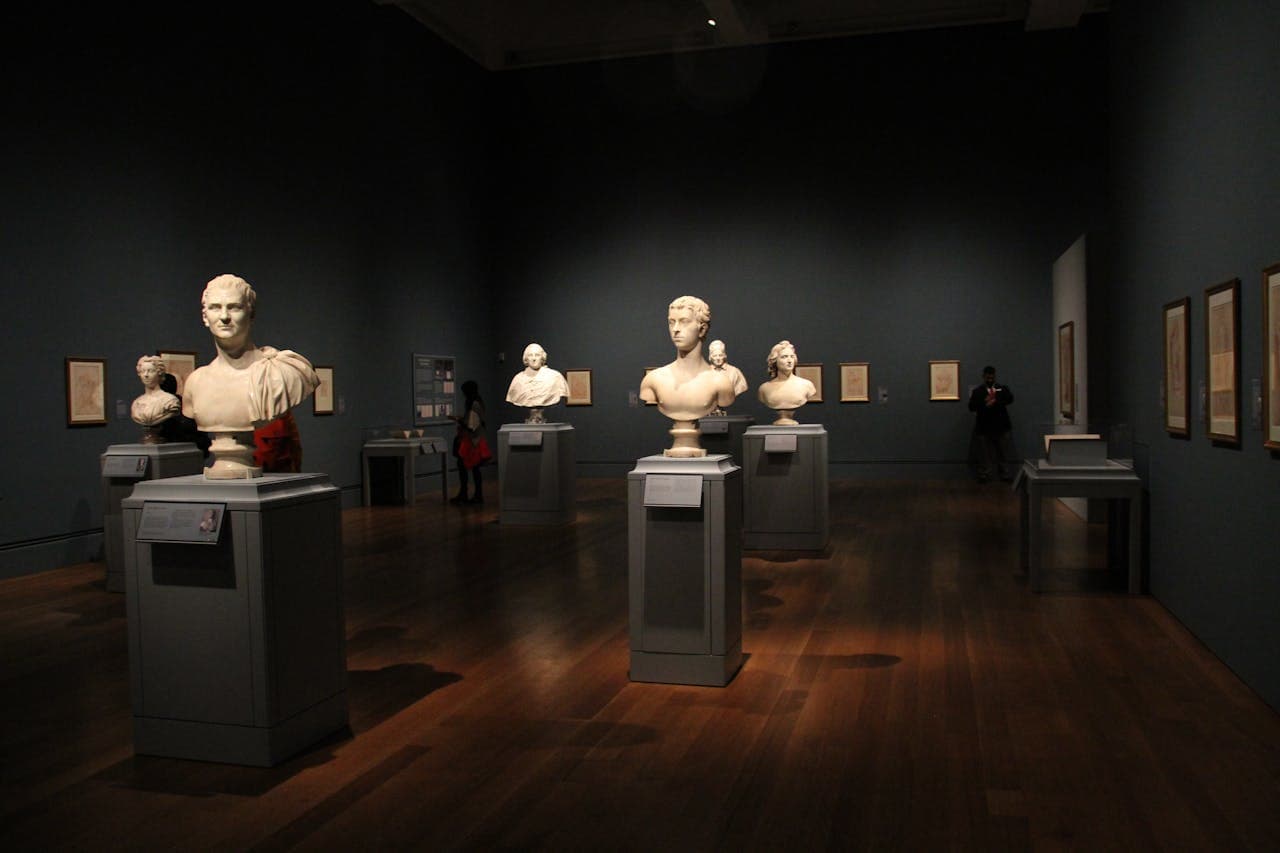 Interior of an art museum gallery displaying classical marble busts on pedestals, with framed artwork on dark blue walls and wooden flooring.