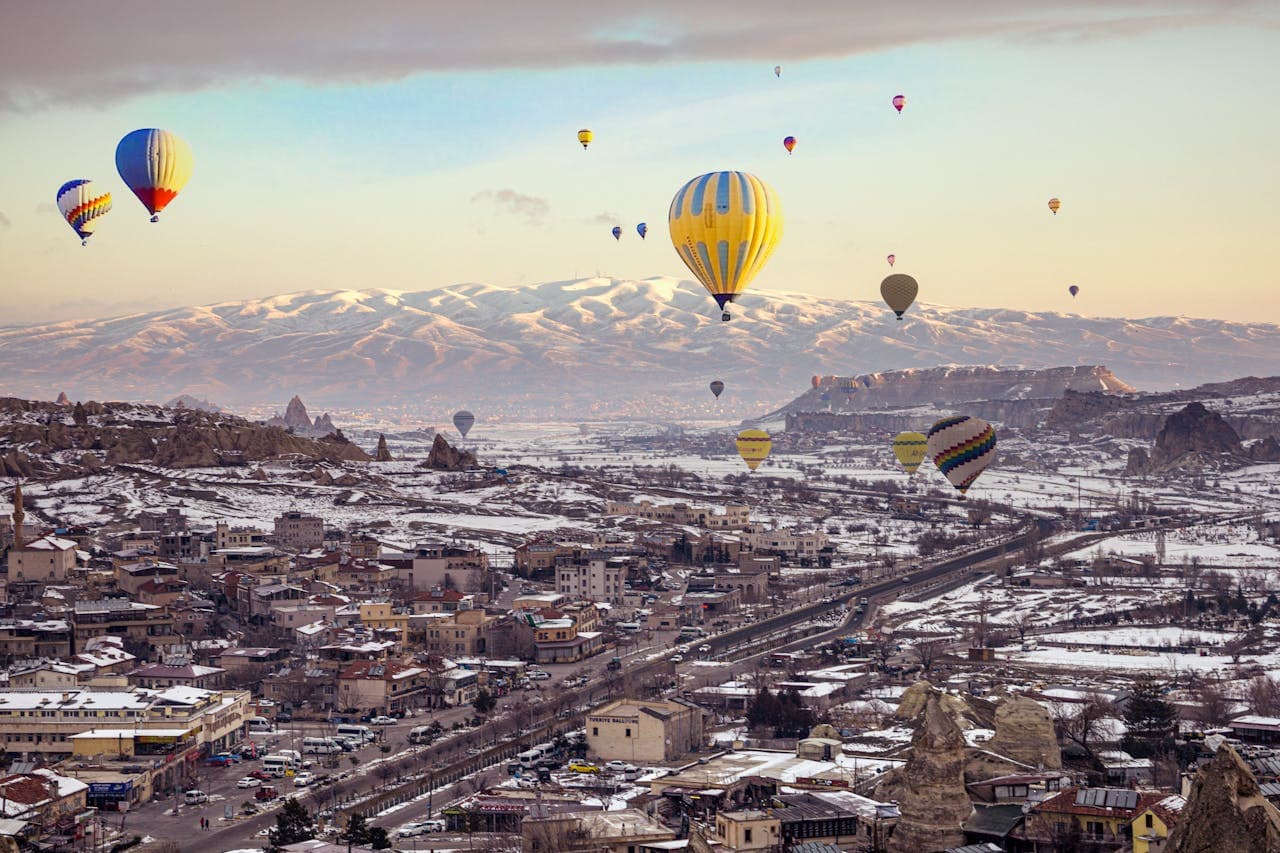 Hot air balloons float over a snowy Cappadocia landscape, with rocky formations and distant mountains under a soft, cloudy sky.