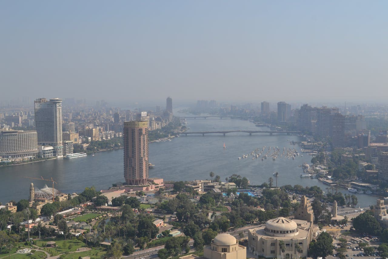 A cityscape featuring the Nile River running through a modern urban area, with high-rise buildings, bridges, and boats on the water, surrounded by greenery and city infrastructure.