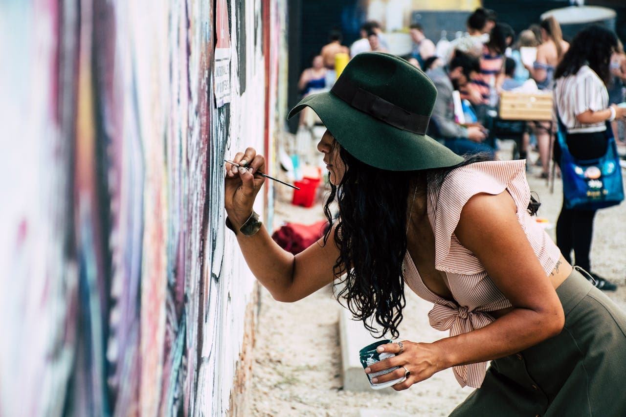 A woman in a wide-brimmed hat and ruffled top painting a mural on a wall at an outdoor art event.