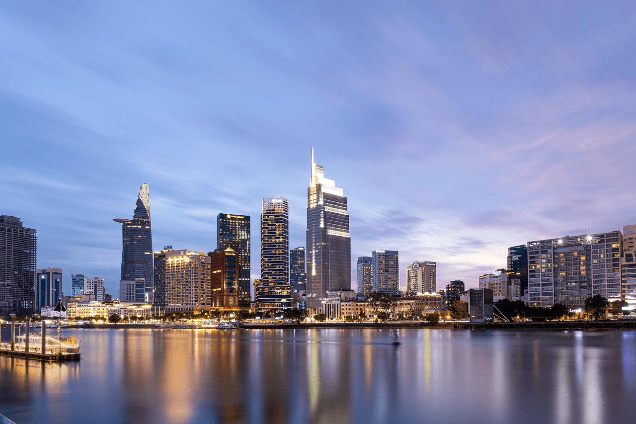 Skyline of a city with modern skyscrapers reflecting on a calm river during twilight, under a cloudy blue and pink sky.