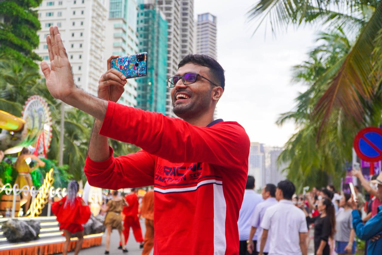 Man in a red shirt taking a selfie with a colorful phone during a lively outdoor parade, with tall buildings, palm trees, and crowds in the background.