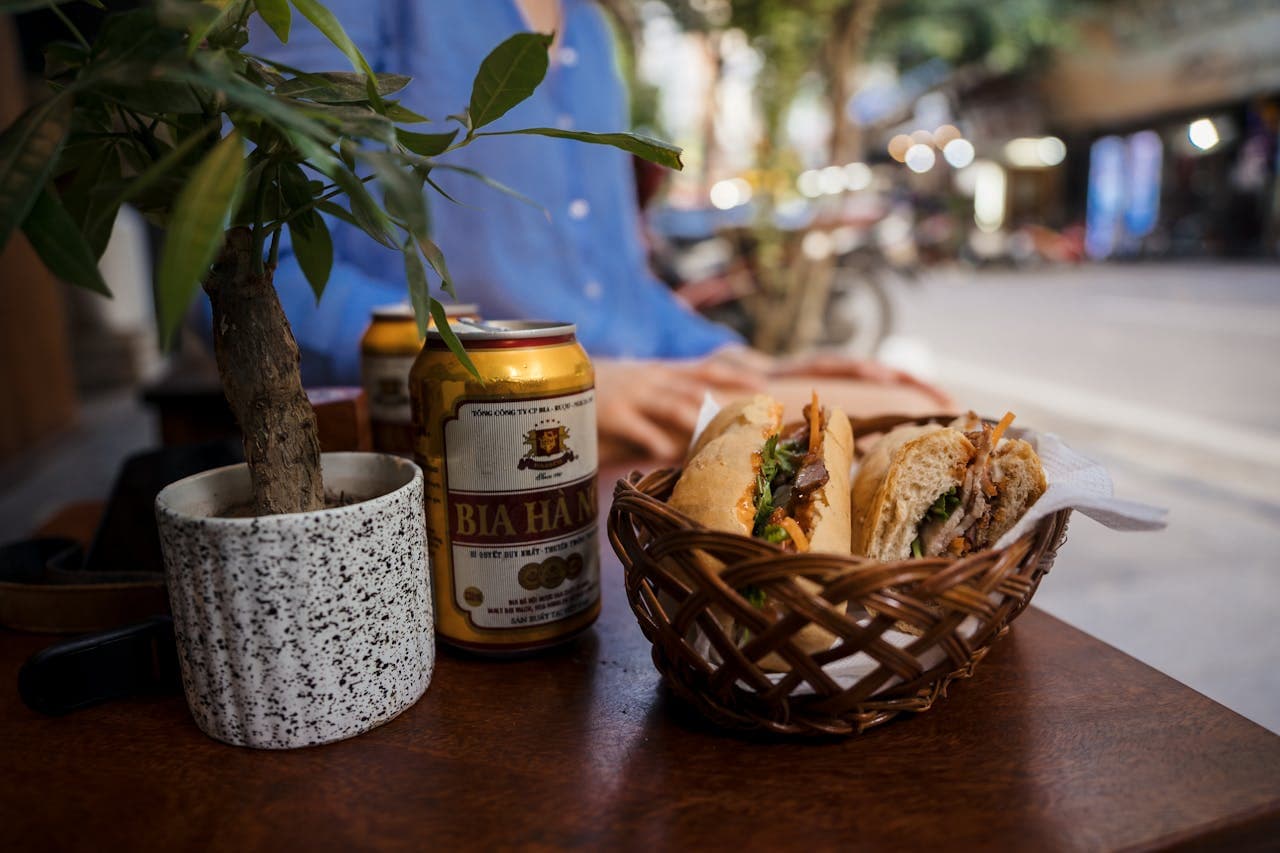 Vietnamese banh mi sandwich in a wicker basket with cans of Bia Ha Noi beer and a potted plant on a wooden table at an outdoor street café.