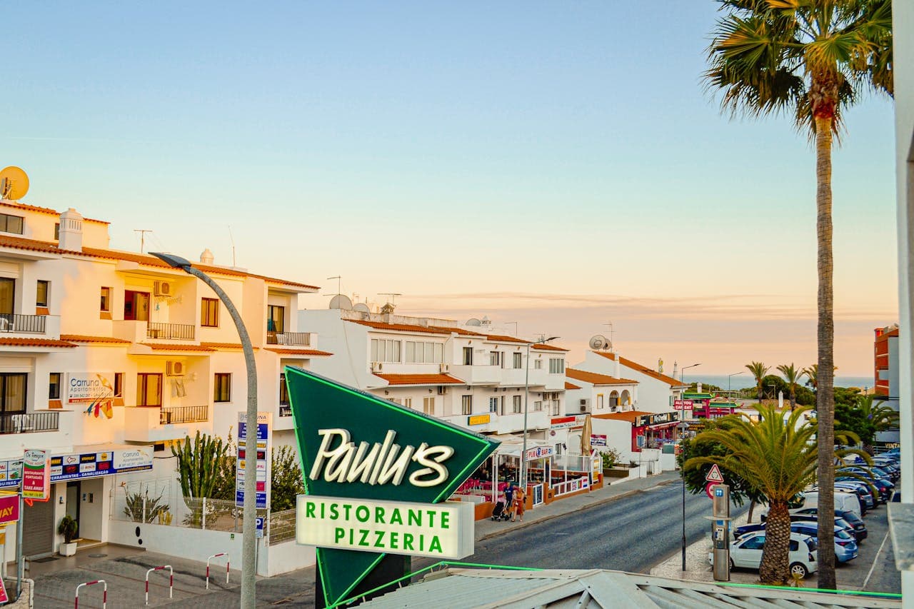 A lively street scene in a Mediterranean town at sunset, featuring white buildings with terracotta roofs, palm trees, parked cars, and a prominent neon sign for "Paulu's Ristorante Pizzeria" in the foreground.